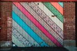 A brick wall covered with large, colorful diagonal stripes. Each stripe features different patterns, including floral, geometric, and ornate designs in vibrant colors.