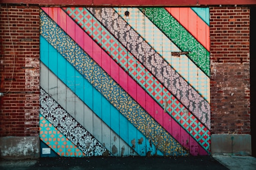 A brick wall covered with large, colorful diagonal stripes. Each stripe features different patterns, including floral, geometric, and ornate designs in vibrant colors.
