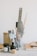 A collection of art and writing tools organized in a transparent cup. Items include pencils, a ruler, brushes, scissors, and a stapler. Nearby, there are bottles of ink, a small block, and stacks of notebooks. The setting is minimalist with muted tones, creating an organized and clean workspace aesthetic.