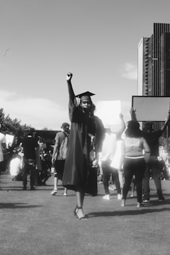 A person in a graduation gown and cap stands with their fist raised as part of a crowd gathered outdoors. The background includes people holding signs, suggesting a demonstration or protest. Tall buildings are visible in the background, and the image is in black and white.