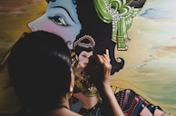 A person with dark hair is painting a detailed portrait that includes a large face profile with a green and pearl headdress. The artist uses fine brush strokes to complete the intricate artwork, which shows traditional attire with vibrant colors.