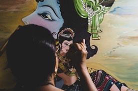 A person with dark hair is painting a detailed portrait that includes a large face profile with a green and pearl headdress. The artist uses fine brush strokes to complete the intricate artwork, which shows traditional attire with vibrant colors.
