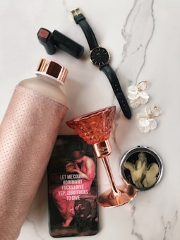A collection of stylish personal items laid out on a marble surface. Features include a leather-covered water bottle with a metallic lid, a black wristwatch with a gold buckle, a pair of gold and pearl drop earrings, a lipstick with black casing labeled 'MATT 20', a pink crystal goblet with a metallic stem, and a compact mirror with a vintage image. Also present is a phone case displaying text and an image of a classical painting.