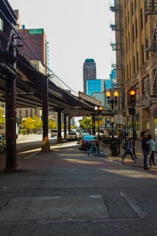 A city street scene features a row of parked cars along the sidewalk under an elevated train track. The street is flanked by tall buildings, including one with visible fire escapes. Pedestrians are visible, including a couple walking near a crossing. Traffic and pedestrian lights are visible, with a sign indicating traffic regulations. Trees are adding greenery to the urban environment.
