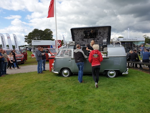 An outdoor event features a retro camper van converted into a mobile bar, attracting several people gathered around. The scene includes other stalls and vehicles in a grassy area, with a cloudy sky above.