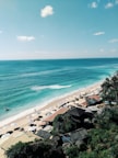 A scenic view of a tranquil beach with turquoise waters and gentle waves lapping at the shore. Sun loungers shaded by white umbrellas line the sandy coastline, while a few people stroll along the water's edge. Rustic beach huts with thatched roofs are nestled among lush greenery along the cliffside, enhancing the tropical vibe.