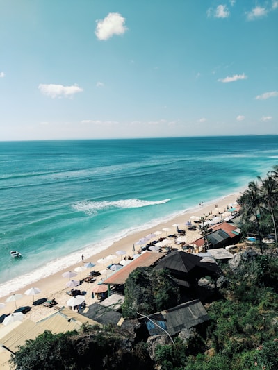A scenic view of a tranquil beach with turquoise waters and gentle waves lapping at the shore. Sun loungers shaded by white umbrellas line the sandy coastline, while a few people stroll along the water's edge. Rustic beach huts with thatched roofs are nestled among lush greenery along the cliffside, enhancing the tropical vibe.