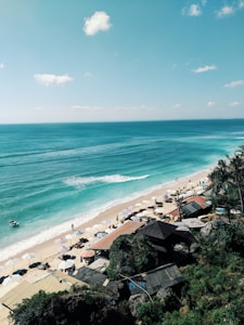 A scenic view of a tranquil beach with turquoise waters and gentle waves lapping at the shore. Sun loungers shaded by white umbrellas line the sandy coastline, while a few people stroll along the water's edge. Rustic beach huts with thatched roofs are nestled among lush greenery along the cliffside, enhancing the tropical vibe.