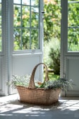 A wicker basket filled with fresh lavender and herbs is placed on a sunlit floor inside an open doorway that leads to a lush garden. Soft shadows and natural light create a serene and inviting atmosphere.