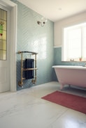 A bathroom features a classic clawfoot bathtub with pink exterior and an elegant brass radiator mounted on a herringbone-patterned, teal-tiled wall. A small window allows natural light to illuminate the space which includes an ornate wall sconce and a stained glass door panel. A textured pink mat lies on the white marble floor, adding warmth to the cool-toned decor.