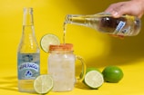 A bottle of sparkling water is placed next to a clear glass mug filled with ice and a lime wedge on the rim. Sparkling water is being poured into the glass from another bottle held by a hand. Two whole limes are positioned around the glass, and the background is a bright yellow.