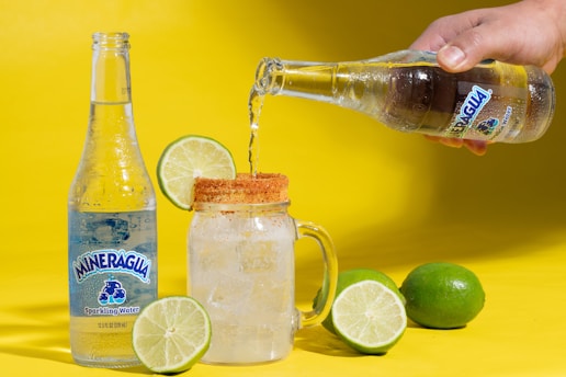 A bottle of sparkling water is placed next to a clear glass mug filled with ice and a lime wedge on the rim. Sparkling water is being poured into the glass from another bottle held by a hand. Two whole limes are positioned around the glass, and the background is a bright yellow.