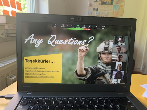 A laptop screen displaying a Zoom call interface with several participants visible on the right side, one of whom is waving. The main part of the screen shows a slide with text 'Any Questions?' and a background image of a person in military attire holding an item. There is a yellow box with contact information in Turkish below the text. The setting appears to be a desk near a window with light coming through.