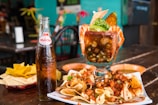 A large glass filled with a seafood cocktail is garnished with shrimp, avocado, and fresh herbs. Surrounding the glass are a variety of chips with mixed nuts and salsa spread on a plate, accompanied by a glass bottle of Sidral Mundet soda. The setting appears to be a rustic restaurant with wooden tables.