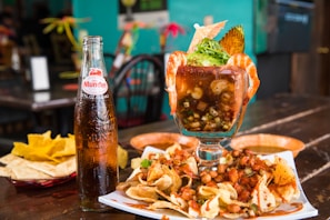 A large glass filled with a seafood cocktail is garnished with shrimp, avocado, and fresh herbs. Surrounding the glass are a variety of chips with mixed nuts and salsa spread on a plate, accompanied by a glass bottle of Sidral Mundet soda. The setting appears to be a rustic restaurant with wooden tables.