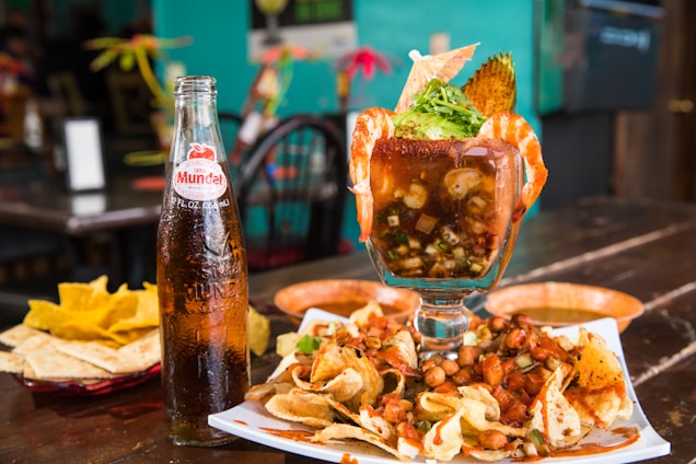 A large glass filled with a seafood cocktail is garnished with shrimp, avocado, and fresh herbs. Surrounding the glass are a variety of chips with mixed nuts and salsa spread on a plate, accompanied by a glass bottle of Sidral Mundet soda. The setting appears to be a rustic restaurant with wooden tables.