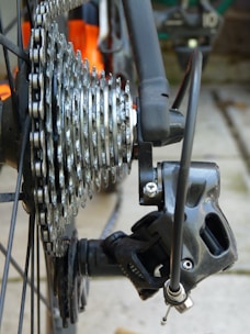 A close-up view of a bicycle gear system. The image features a metallic gear cassette with multiple cogs and a derailleur. The shiny metal surfaces exhibit signs of use, with some dirt visible on the components. The background shows a blurred outdoor setting.