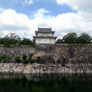 The image features a traditional Japanese castle structure situated atop a large stone wall. The wall, made of large, gray and beige stones, reflects in a calm body of water in the foreground. Lush green trees surround the building, and the sky above is partly cloudy, providing a serene backdrop.