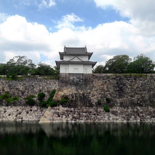The image features a traditional Japanese castle structure situated atop a large stone wall. The wall, made of large, gray and beige stones, reflects in a calm body of water in the foreground. Lush green trees surround the building, and the sky above is partly cloudy, providing a serene backdrop.