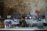 A cozy indoor setting with a rustic stone wall adorned with various artworks, photographs, and decorative items. There is a man standing behind a counter surrounded by books, glasses, and other items. A vase with purple flowers is placed on the counter, along with some brochures and a Cyprus guidebook.