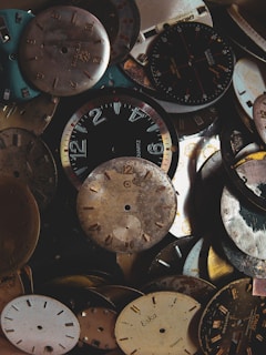 A collection of assorted vintage clock faces and watch dials, some with visible brands and numbers. The dials are stacked and overlapping each other, with a mix of worn and polished metal surfaces showcasing a range of colors and textures.