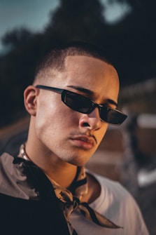 A person wearing sunglasses and a stylish jacket is looking confidently at the camera. The image is taken in low light, adding dramatic shadows across the face. The background is blurred, highlighting the subject's facial features and accessories.
