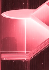 A geometric architectural structure with strong angular lines and a pink tint. The design features layered, horizontal panels and an inset lighting element that creates contrast against the darker sections.