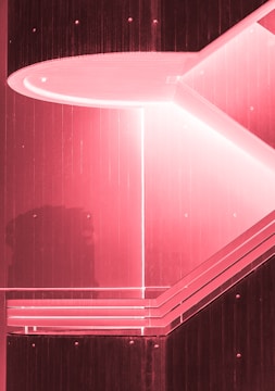 A geometric architectural structure with strong angular lines and a pink tint. The design features layered, horizontal panels and an inset lighting element that creates contrast against the darker sections.