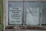 Two paper notices are taped to a window. The one on the left promotes the 1st free black school building in the Northwest Territory, noting an open house event on the third Sunday in September. The notice on the right lists contact information for arranging visits at other times, including names and phone numbers.