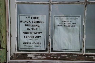 Two paper notices are taped to a window. The one on the left promotes the 1st free black school building in the Northwest Territory, noting an open house event on the third Sunday in September. The notice on the right lists contact information for arranging visits at other times, including names and phone numbers.