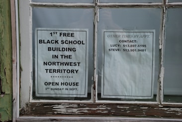 Two paper notices are taped to a window. The one on the left promotes the 1st free black school building in the Northwest Territory, noting an open house event on the third Sunday in September. The notice on the right lists contact information for arranging visits at other times, including names and phone numbers.