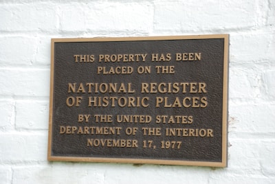 A metal plaque is mounted on a white brick wall, displaying the text indicating the property's inclusion on the National Register of Historic Places. The wording states the designation by the United States Department of the Interior with the date November 17, 1977.