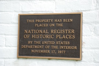 A metal plaque is mounted on a white brick wall, displaying the text indicating the property's inclusion on the National Register of Historic Places. The wording states the designation by the United States Department of the Interior with the date November 17, 1977.