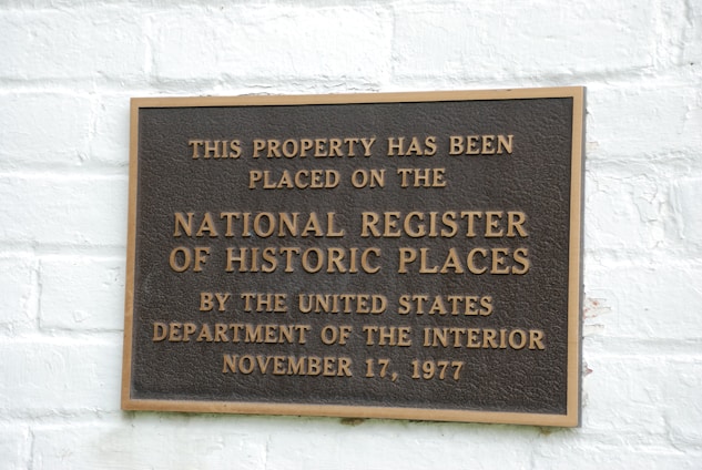 A metal plaque is mounted on a white brick wall, displaying the text indicating the property's inclusion on the National Register of Historic Places. The wording states the designation by the United States Department of the Interior with the date November 17, 1977.