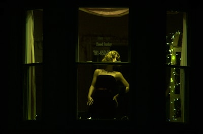 A mannequin is seen in a dark window display, illuminated by artificial green light. The mannequin appears to be wearing a strapless dress and has styled hair. The background features soft, decorative lights and partially visible signage.