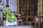 A person in a green robe is seated on a chair, holding their face with one hand, inside a building with ornate stone walls and a metal gate. The person appears to be deep in thought or prayer.