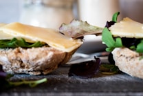 Slices of bread topped with fresh greens and slices of cheese are arranged closely. The image focuses on the texture of the bread and the freshness of the ingredients, creating an impression of a rustic and appetizing sandwich preparation. In the background, there is a soft focus on jars or containers, adding a homely feel.