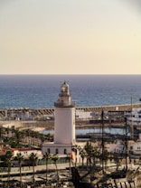 An ocean view with a white lighthouse positioned prominently in the foreground. Surrounding the lighthouse are palm trees and several boats docked in the marina. A historic ship with tall masts is also visible. The sky is clear, and the sea stretches out in the background, creating a peaceful maritime atmosphere.