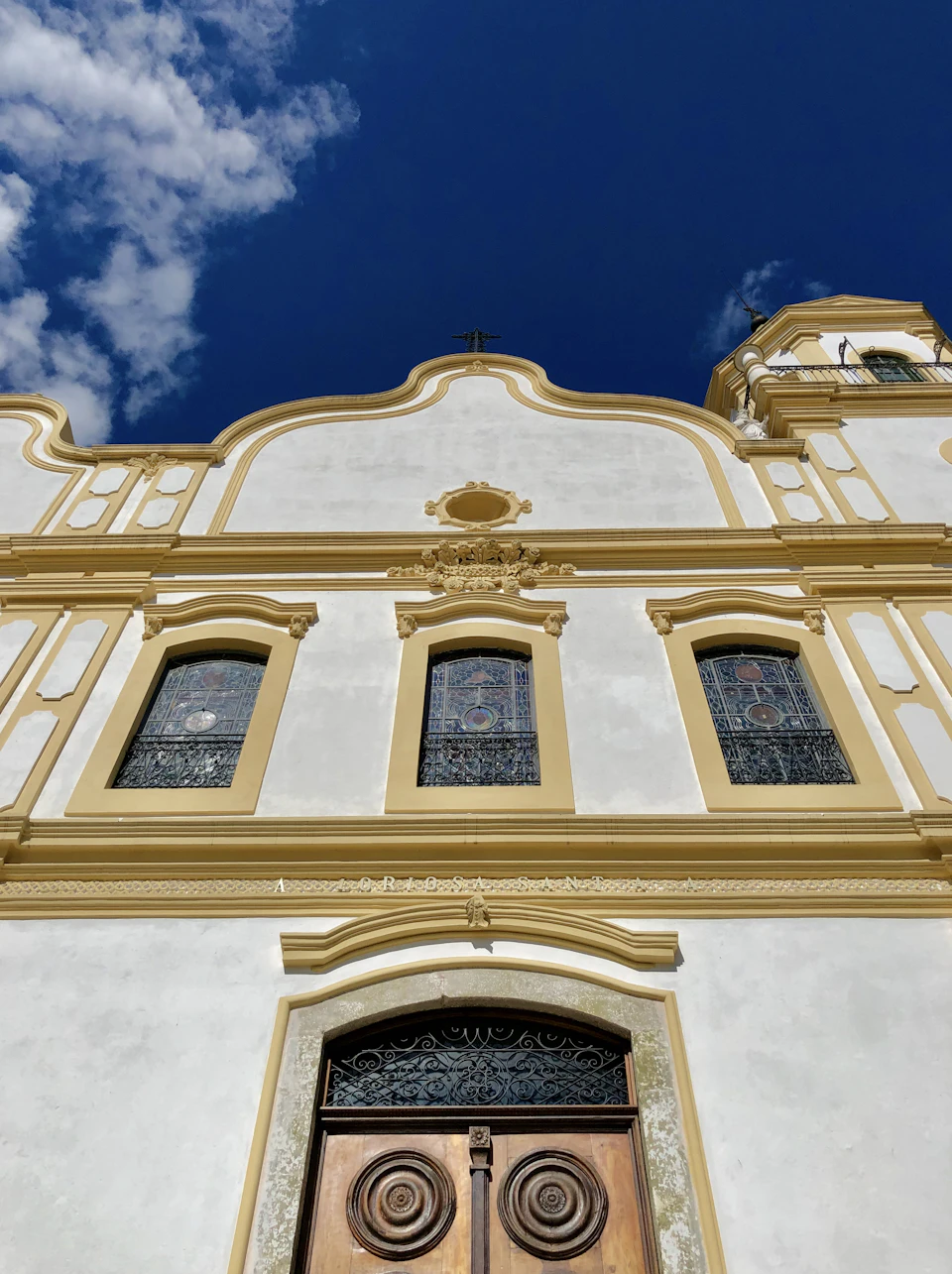 The image shows a white building with yellow trim, featuring three windows on each side and a large archway at the entrance. The building has a pointed roof and is topped by a cross, suggesting it may be a church or other religious structure. The sky above is blue with scattered clouds, providing a serene backdrop to the architectural details of the building.