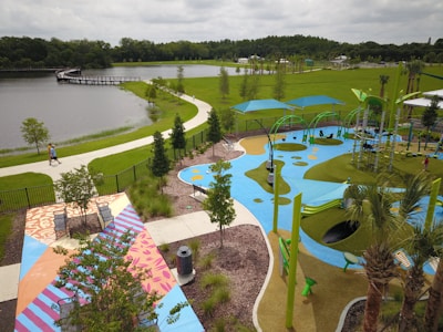 A vibrant outdoor park featuring a colorful playground with blue, green, and yellow play surfaces. There are modern playground equipment including swings and climbing structures under large shade canopies. A pathway winds along a lake surrounded by lush green grass and trees. The area includes benches and decorative patterned pavement near landscaped garden beds.