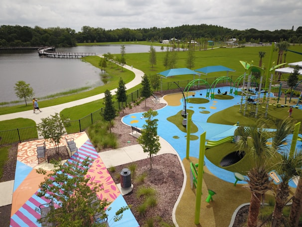 A vibrant outdoor park featuring a colorful playground with blue, green, and yellow play surfaces. There are modern playground equipment including swings and climbing structures under large shade canopies. A pathway winds along a lake surrounded by lush green grass and trees. The area includes benches and decorative patterned pavement near landscaped garden beds.