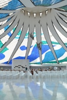A large, open interior space features a striking stained glass ceiling with blue, green, and white geometric patterns. Angel sculptures hang from the ceiling, and rows of white chairs are arranged below on the polished floor. The ambiance is serene and spacious, with a few people visible in the background.