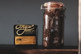 A clear glass jar filled with roasted coffee beans is placed next to a black package of Onyx Coffee Lab's Ethiopia Agaro Family coffee. The package contains detailed information about the coffee, including its origin and flavor notes. The background is a plain, gray wall, and the items are positioned on a dark wooden surface.