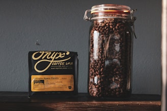 A clear glass jar filled with roasted coffee beans is placed next to a black package of Onyx Coffee Lab's Ethiopia Agaro Family coffee. The package contains detailed information about the coffee, including its origin and flavor notes. The background is a plain, gray wall, and the items are positioned on a dark wooden surface.