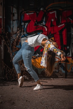 A person is performing a dynamic dance move with their back arched and arms extended. They are dressed in casual attire, including a white lacy top and blue jeans. The background features a vibrant and colorful graffiti wall, adding an urban and artistic atmosphere.