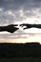 Two hands reaching toward each other in silhouette against a cloudy sky