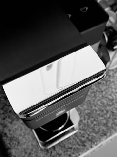 A sleek, modern espresso machine featuring a glossy metallic finish and a black body is placed on a speckled countertop. The surface reflects light, emphasizing its polished appearance. In the background, there's a row of circular objects, possibly coffee pods.