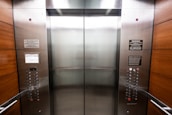 An elevator interior with sleek, metallic doors and wood panel siding. Control panels with buttons and warning signs are situated on either side, with clear labels and numbers. The lighting is bright, accentuating the polished surfaces.