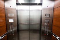 An elevator interior with sleek, metallic doors and wood panel siding. Control panels with buttons and warning signs are situated on either side, with clear labels and numbers. The lighting is bright, accentuating the polished surfaces.