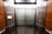 An elevator interior with sleek, metallic doors and wood panel siding. Control panels with buttons and warning signs are situated on either side, with clear labels and numbers. The lighting is bright, accentuating the polished surfaces.